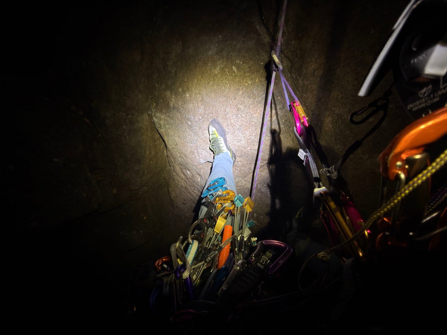 A climbing shoe on the rock and a trad rack of cams and nuts, lit by a headlamp at night, with a purple rope running up the wall